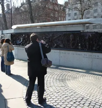 The Battle of Britain Monument with a silver sculpture above dark relief panels and carved stone text, with people standing nearby.