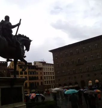 Cosimo I Equestrian Monument seen from behind in a wet Florence square with umbrellas and a Generali building in the background.
