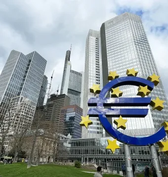 Blue euro sculpture with yellow stars in front of Frankfurt skyscrapers and a cloudy sky.