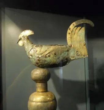 Gallo di Ramperto, a weathered metallic bird sculpture with traces of gold on its crest and tail, mounted on a tiered spherical pedestal inside a display case.
