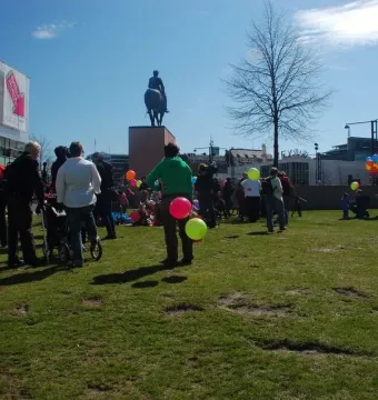 Marshal Mannerheim equestrian statue seen from behind on a tall plinth in a grassy park, with people gathered around and colorful balloons nearby.