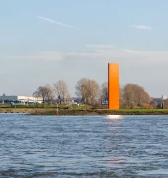 Rheinorange sculpture standing beside a wide river with sunlight reflecting on the water and distant buildings in the background.