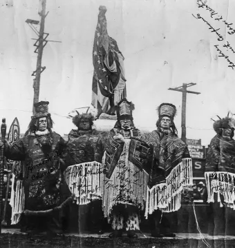 Six men in ceremonial attire stand near the Chief Seattle statue dedication site beside a flagpole.