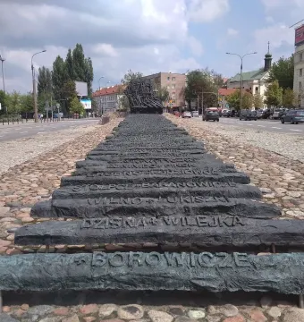 Monument to the Fallen and Murdered in the East stretching along a cobblestone median in Warsaw, with dark inscribed bars leading into a jagged structure.