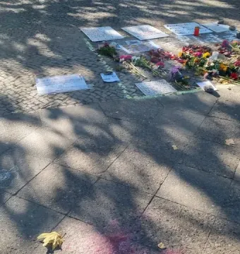 Flowers, signs, and a red candle laid on cobblestones within a green chalk outline near the Peace Statue.