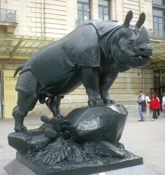 Large rhinoceros sculpture with a textured hide standing on a decorated base outdoors near a stone building.