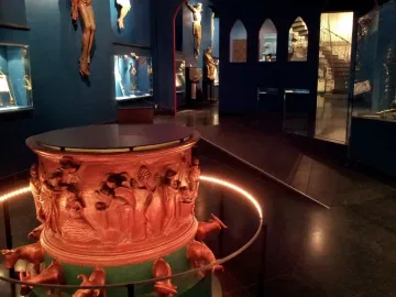 Baptismal font with reddish-brown sculpted reliefs and small animal figures around the base, displayed in a dim gallery.