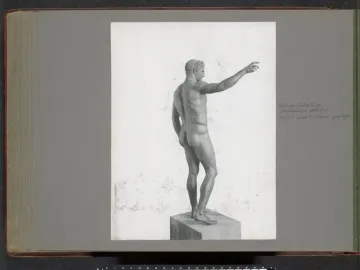 Rear-left view of the Antikythera Ephebe bronze statue, showing a nude male figure on a plinth with one arm extended.