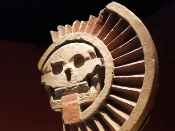 Disk of Mictlāntēcutli stone sculpture with a skull-like face and semicircular ribbed frame in a museum display.
