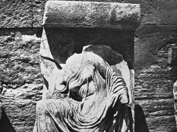 Damaged relief sculpture of Nike Untying Her Sandal, shown headless and draped in flowing robes beside a stone wall.
