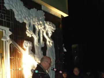 Plon sculptural relief mounted on a brick wall, brightly lit with people gathered below.