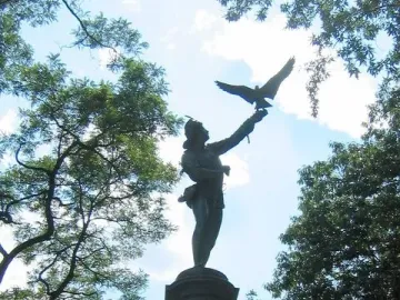 The Falconer statue silhouetted against a bright sky in Central Park, with trees behind it.