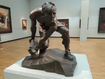 Bronze sculpture of a crouching man holding a rough stone, displayed on a pedestal in a gallery.