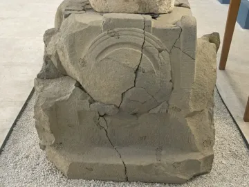 Weathered stone block with carved semicircular motifs and faint inscription lines, showing heavy cracking and damage.