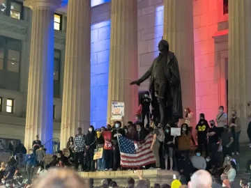 A George Washington statue stands between stone pillars as a crowd gathers below with signs and an American flag.