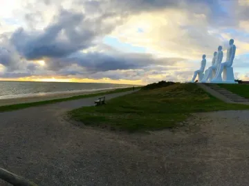 The Man by the Sea sculpture overlooking a beach and the sea at sunset, with four white seated figures on a grassy mound.