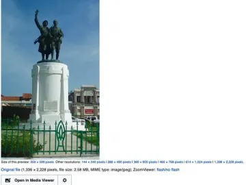 Monument Demba et Dupont with two dark figures on a tall white cylindrical base behind a green fence.