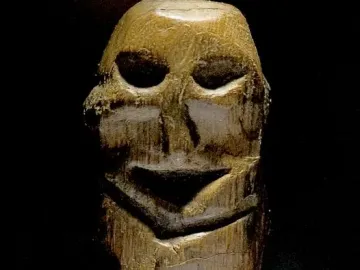Mannetje van Willemstad wooden figure with a weathered surface and hollowed facial features on a clear base.