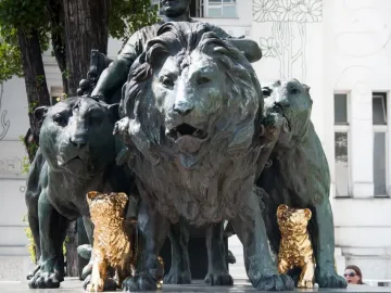 Marc Anton Group bronze sculpture with large lions and two small golden cub figures at the base, outdoors near trees and a white building.