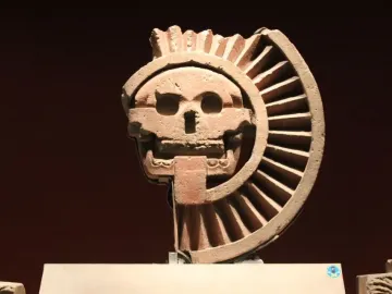 Disk of Mictlāntēcutli showing a skull-like face with hollow eyes and teeth, framed by a ridged semicircular form.