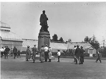 William Henry Seward statue on a tiered pedestal in Volunteer Park, with people gathered nearby and glass greenhouses in the background.