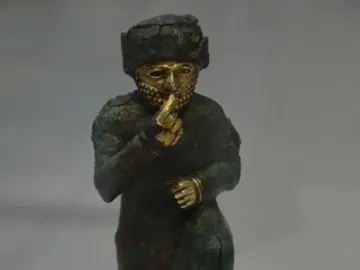 Worshipper of Larsa kneeling with one hand raised to its mouth, showing a dark bronze body and a golden face and beard.