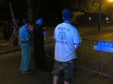 People stand near crowd control barriers at night beside a N.O.P.D. police line sign, with one person wearing a \