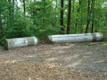 Two weathered Capitol Columns stone segments lie on a dirt and woodchip path surrounded by dense green foliage.