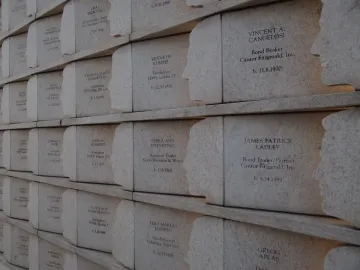 The Postcards memorial shows rows of engraved name plaques on a light stone wall, each with a carved human silhouette along the edge.