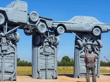 Carhenge made from grey-painted cars and trucks standing in a grassy field under a clear blue sky, with a person looking up at the structure.