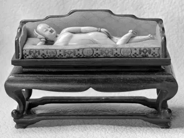 Doctor\'s Lady, a light-colored carved female figure reclining on a patterned daybed with an upholstered back.