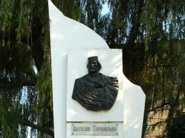 Garibaldi Monument with a dark bust relief and plaques on a white angular pedestal, surrounded by greenery.
