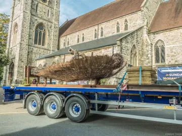 The Knife Angel sculpture леж on a blue flatbed trailer beside a road, with a stone church and clock tower in the background.
