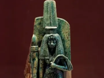 Greenish-blue statue of Queen Tiye wearing a tall ornate headdress and long textured garment, holding objects in both hands.
