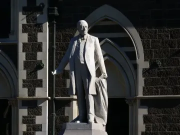 Rolleston Statue of a formally dressed man holding draped fabric beside a dark stone building.