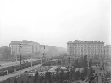 A large Stalin monument plinth stands in a broad square beside an avenue lined with crowds and administrative buildings.