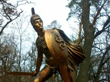 Bronze Greek warrior monument showing a helmeted soldier with a shield and raised sword.