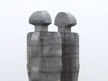 Two colossal abstract concrete figures of the Monument to Humanity stand on a hillside with scaffolding between them and two people at the base.