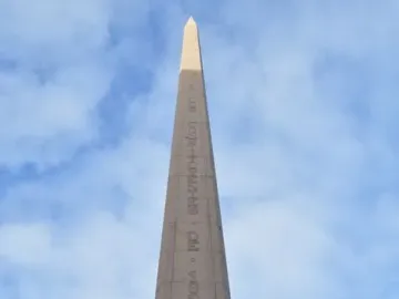 The Obelisk of Montevideo rises against a blue sky, with bronze figures at its base.