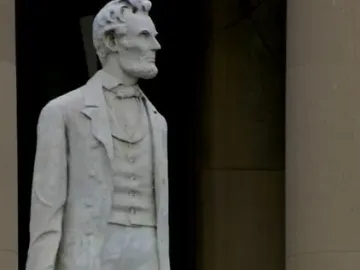 Marble statue of Abraham Lincoln standing in a suit with one hand resting on a bundle of rods.