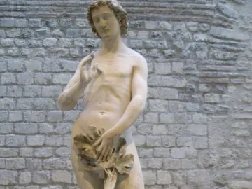 Adam, a slender marble statue with curly hair and foliage covering the groin, standing against a stone wall.
