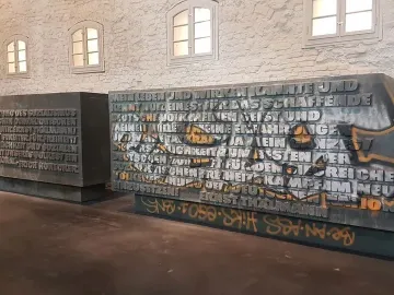 Two inscribed plaques from the Ernst Thälmann Memorial, one marked with orange graffiti, against a brick wall with arched windows.