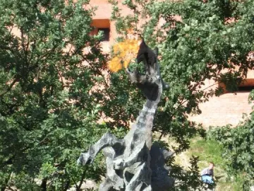 Wawel Dragon sculpture with a flame coming from its open mouth, surrounded by greenery and a brick building in the background.