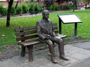 Alan Turing Memorial bronze statue of a man sitting on a park bench holding an apple, with an information board nearby.