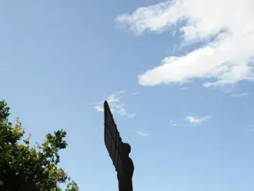 The Angel of the North sculpture stands on a grassy hill beside a paved path with people walking toward it under a blue sky.