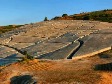 Il Grande Cretto’s cracked concrete slabs spread across a hillside beneath wind turbines and a blue sky.
