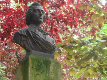 Dark metallic bust of Frédéric Chopin mounted on a weathered stone block amid red and green foliage.