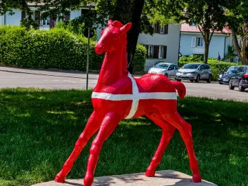 Holbeinpferd sculpture painted bright red with a white cross, standing on a concrete base in a grassy area.