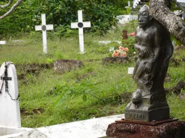 Oviri sculpture, a dark figure holding a smaller creature, standing in a grassy cemetery.