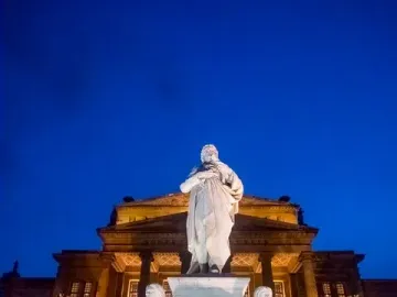 Schiller Monument illuminated at night in front of the Konzerthaus in Berlin, with seated allegorical figures beside the central statue.
